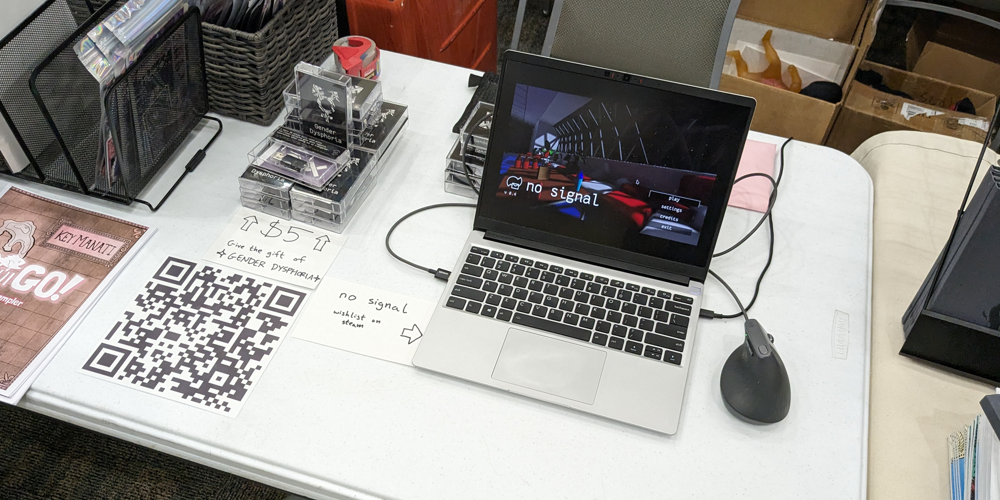 My half of the table at STAPLE! Austin 2024. The table is not covered by a tablecloth. A stack of Gender Dysphoria cassette cases sit to the left, with an index card below it which reads “Give the gift of ⭐ GENDER DYSPHORIA ⭐”. A Framework laptop and a wired ergonomic mouse sits to the right, with an index card pointing at it which reads “no signal” and “wishlist on steam”