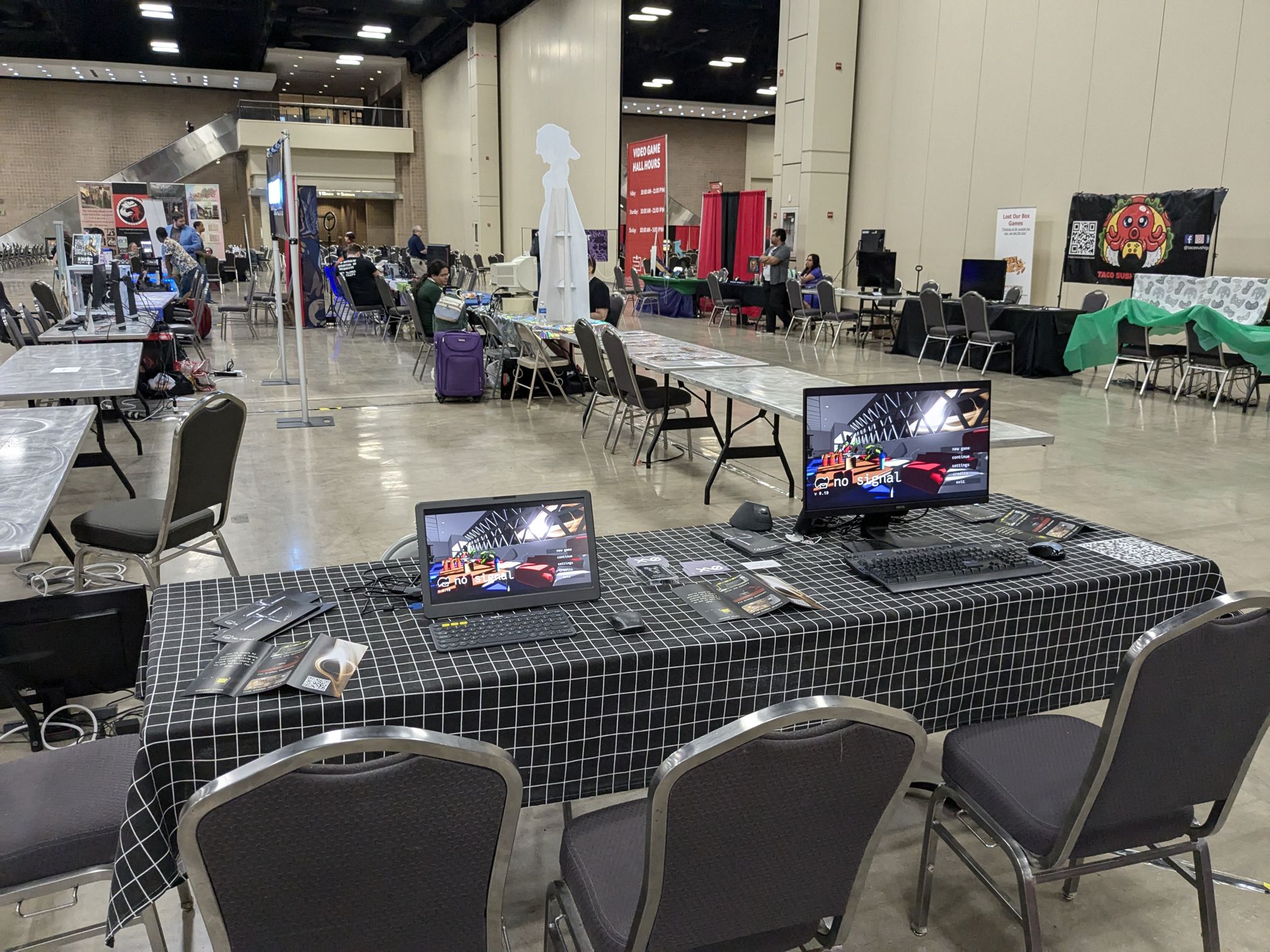 My table at San Japan 2024. It’s covered with a black tablecloth with a white grid pattern. On the table sits two monitors, one of which is portable and the other is not. There are also flyers on the table.