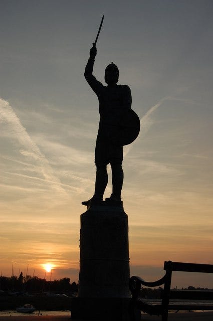 A statue of an armed man, sword raised, silhouetted against the sunset.