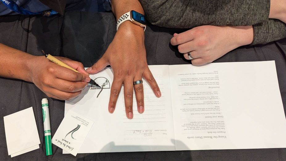 Two people playing The Bonsai Diary. A pair of hands on the left is using a brush pen to draw a tree, while the pair of hands on the right are folded, waiting.