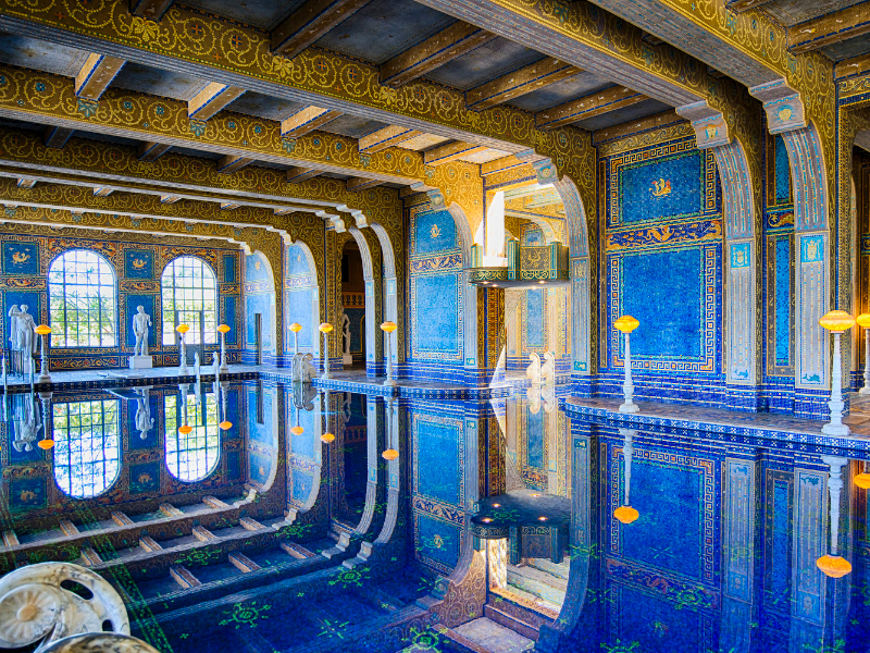 The Roman Pool at Hearst Castle, an elaborately decorated indoor pool with a blue and gold aesthetic. The water is completely still, causing a perfect reflection.
