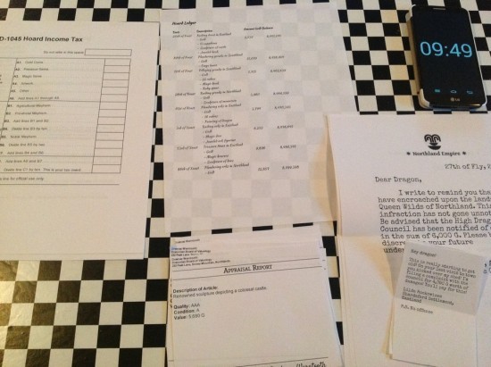 Printed out pages of dragon tax documents organized on a table, in the same layout as the computer game, next to a smartphone displaying 9:49.