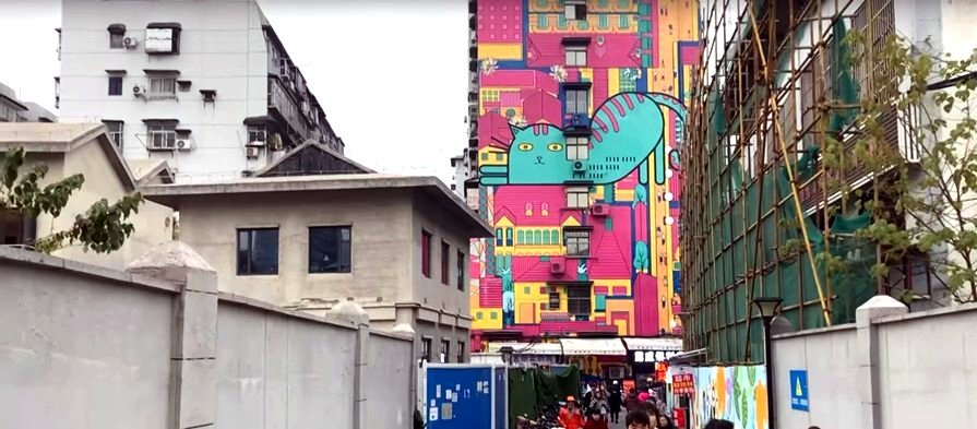 Building painted with a colorful mural of a blue cat, nestled among white and grey buildings at the sides of a crowded street. Building painted with a colorful mural of a blue cat, nestled among white and grey buildings at the sides of a crowded street.