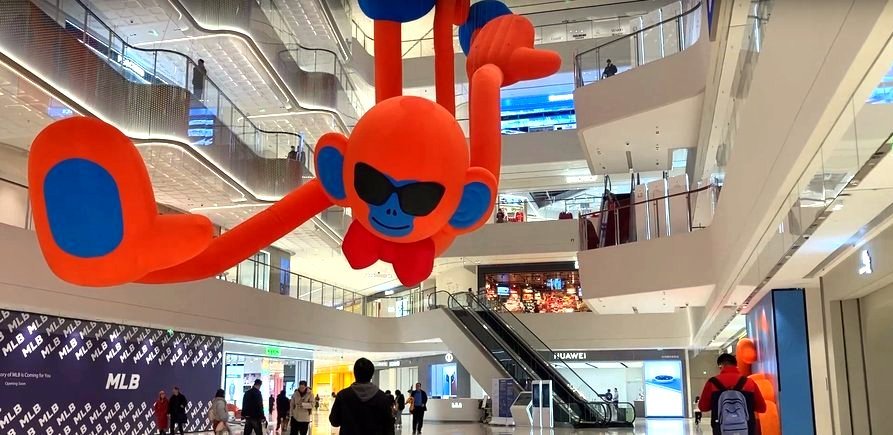 A giant inflatable blue-and-orange monkey with cartoon glasses, hanging from the ceiling of a multi-story shopping mall full of glass, steel, and florescent white light. A giant inflatable blue-and-orange monkey with cartoon glasses, hanging from the ceiling of a multi-story shopping mall full of glass, steel, and florescent white light.