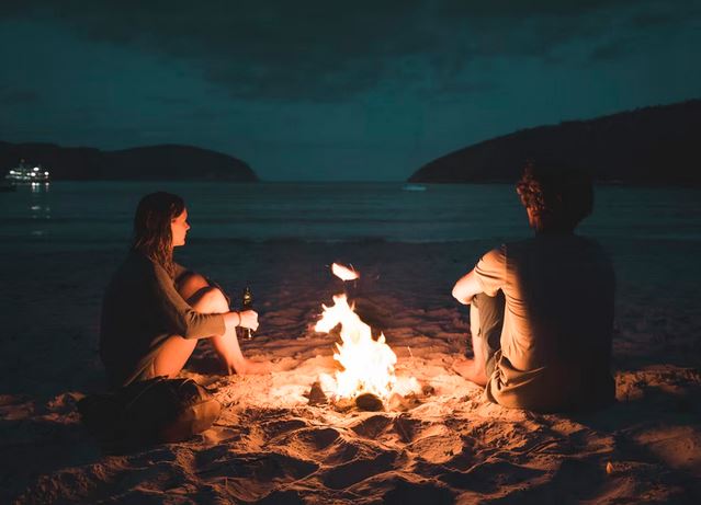 young man and woman near bonfire looking at sea